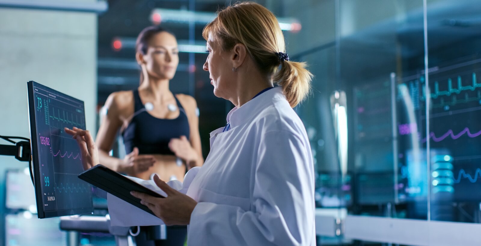 sport scientist supervises touches display showing ekg status while in the background woman athlete running on a treadmill
