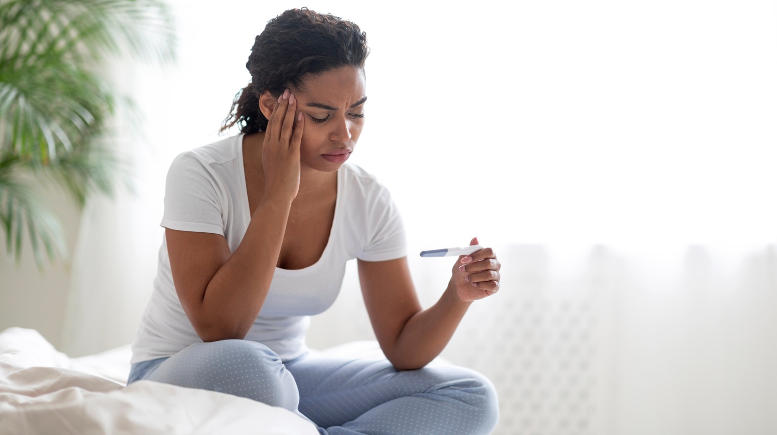 young african american woman with pregnancy test in hand sitting on bed in white bedroom interior